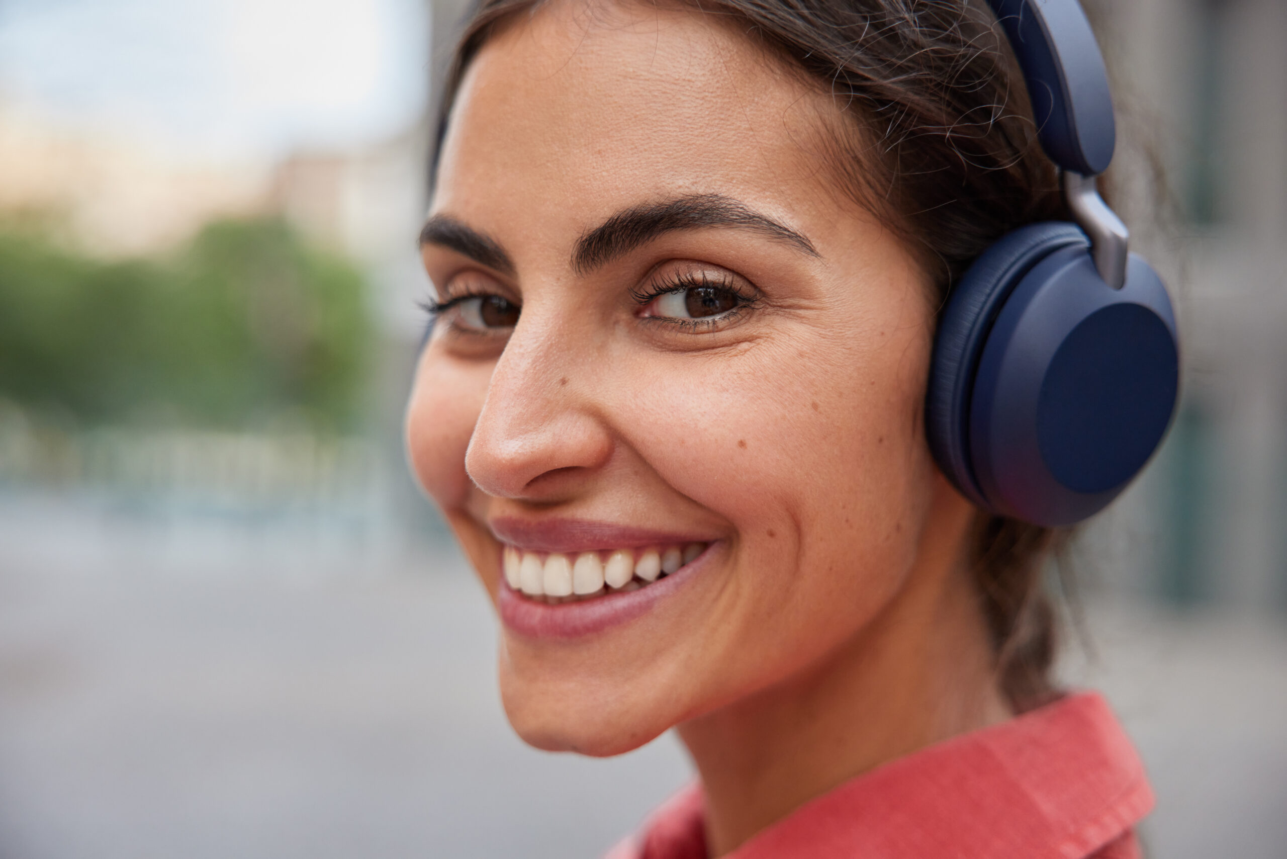 Close up shot of happy young woman smiles toothily enjoys leisure time listens favorite music in wireless headphones poses outdoors against blurred background. People hobby and lifestyle concept